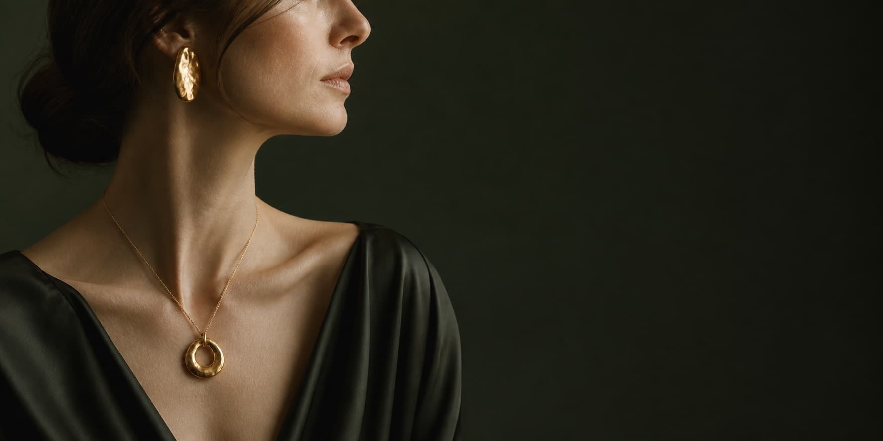Model wearing ÉLORA pendant and organic gold earring against a dark green backdrop