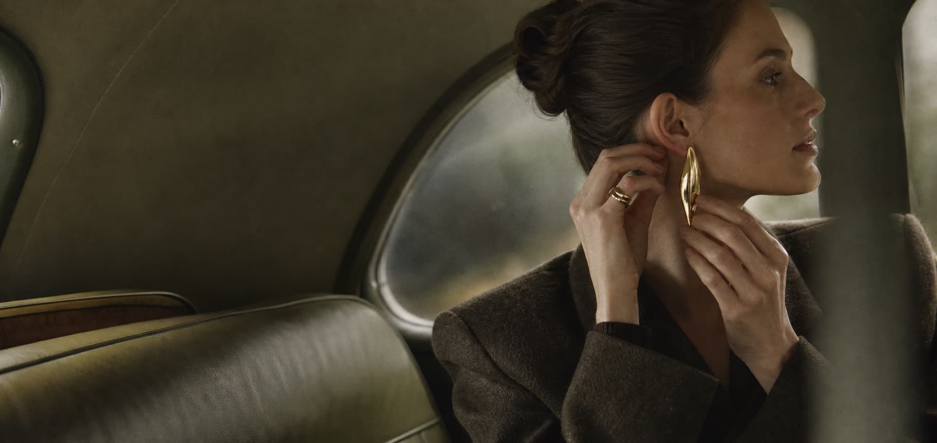 Woman fastening a sculptural gold earring in an olive-toned vintage car
